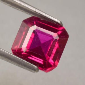 May include: A rectangular-cut ruby gemstone held by silver tweezers. The ruby has a deep red colour with a lighter, almost pink, centre. The gem's facets reflect light, creating a sparkling effect.