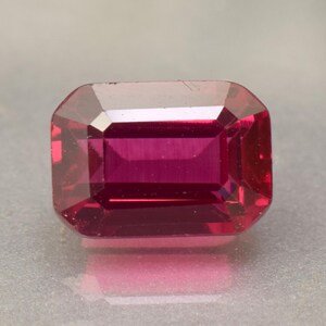 May include: A rectangular-cut gemstone with a deep magenta colour. The gem has a polished surface and a symmetrical shape, reflecting light. The gemstone is likely a ruby or similar precious stone, suitable for jewellery.