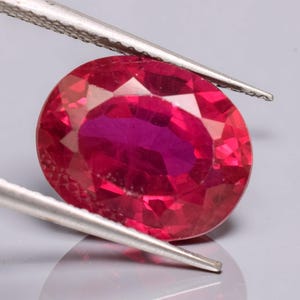 May include: A vibrant, oval-cut ruby gemstone with a deep red hue and multifaceted surface, held by a pair of silver-toned metal tweezers. The gemstone displays internal clarity and light reflection across its polished, symmetrical facets.