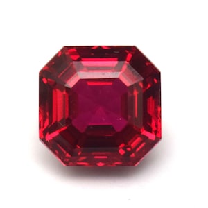 May include: A faceted, square-cut ruby gemstone. The gem is a deep, saturated red color with a brilliant, reflective surface. The cut features multiple facets that catch and reflect light, creating a sparkling effect. The gemstone is set against a white background.