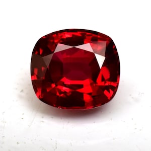 May include: A deep red gemstone with a cushion cut. The gemstone is faceted and has a high polish.