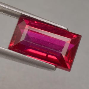 Puede incluir: Una gema de rubí rectangular facetada, sujeta por unas pinzas metálicas. El rubí es de color rojo intenso con una superficie vibrante y reflectante. El corte de la gema muestra su estructura interna y brillo.