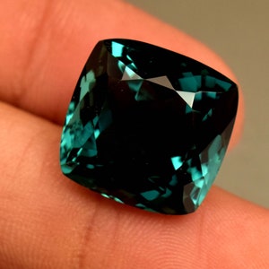 Puede incluir: Una piedra preciosa verde oscuro con talla cojín. La piedra preciosa está facetada y tiene un alto nivel de brillo.