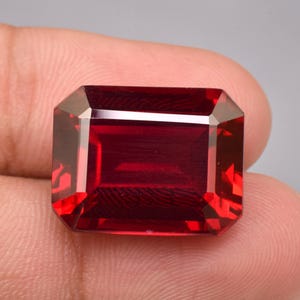 May include: A deep red, rectangular gemstone with an emerald cut, featuring sharp, clean facets that reflect light across its surface. The stone displays a rich, saturated crimson hue with high clarity and a polished, smooth finish throughout.