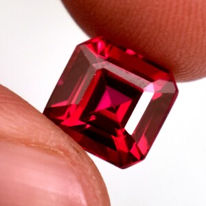 May include: A faceted, rectangular-cut, deep red gemstone. The gem is held between two fingers against a white background. The gemstone's facets reflect light, creating a brilliant display of colour and sparkle. The gem is a vibrant shade of red.
