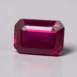 May include: A faceted, rectangular ruby gemstone with a deep red hue. The gem has an emerald cut with beveled edges, reflecting light and showcasing its clarity. The gemstone is set against a neutral background.
