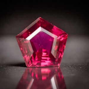 May include: A faceted, pentagon-shaped gemstone with a deep magenta hue. The gem's polished surfaces reflect light, creating a brilliant display of color and facets. The gemstone is set against a dark background, highlighting its vibrant color.