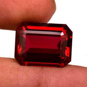 May include: A faceted, rectangular gemstone with a deep red hue. The gem is held between fingers, showcasing its clarity and brilliance. The cut features a series of flat facets, reflecting light and enhancing the stone's color. The gem is likely a ruby or garnet.