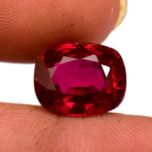 Op de afbeelding: Een gefacetteerde robijn edelsteen met kussenslijpsel. De edelsteen heeft een diepe, verzadigde rode kleur met een hoge mate van helderheid. De robijn wordt tussen twee vingers gehouden, wat de schittering en de slijpvorm laat zien. De edelsteen staat tegen een witte achtergrond.