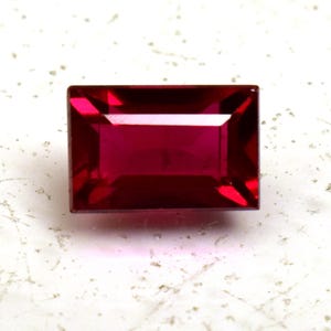 Puede incluir: Una gema de rubí rectangular y facetada, de color rojo intenso. La gema tiene un aspecto limpio y pulido con bordes afilados y una forma simétrica. La piedra está sobre un fondo blanco moteado.