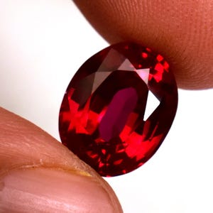 May include: A faceted, oval-shaped ruby gemstone. The gem is a deep, vibrant red color with multiple facets that reflect light. The ruby is held between two fingers against a white background.