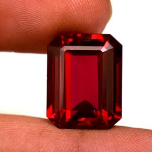 Puede incluir: Una gema roja oscura, tallada en forma rectangular y facetada. La gema tiene una superficie pulida con bordes afilados y limpios. El color es un rojo intenso y saturado, con un ligero brillo interno. La gema se sostiene entre dos dedos.