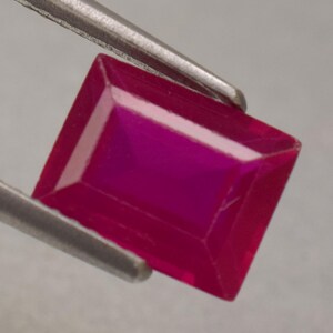 May include: A rectangular-cut ruby gemstone, held by tweezers. The gem is a deep red colour with a polished surface and sharp edges. The facets of the gemstone reflect light, creating a brilliant display. The tweezers are silver.