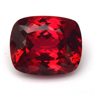 May include: A faceted, cushion-cut gemstone with a deep red hue. The gem's surface reflects light, showcasing its intricate internal structure. The shape is a square with rounded corners, and the overall appearance is vibrant and eye-catching.