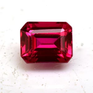 Peut inclure: Une pierre précieuse rubis taillée en forme rectangulaire. La gemme est d'un rouge profond et vibrant, avec une grande clarté, reflétant la lumière. La taille présente une série de facettes parallèles, créant un éclat brillant. La pierre est posée sur un fond blanc.