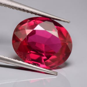 Op de afbeelding: Een levendige, ovaal geslepen robijn met een dieprode kleur en meerdere facetten die het licht reflecteren. De edelsteen wordt vastgehouden door een zilverkleurige metalen pincet, wat de helderheid en het gepolijste oppervlak benadrukt.