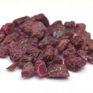 Op de afbeelding: Een stapel ruwe robijn edelstenen, met een dieprode tint met vleugjes bruin en goud. De edelstenen zijn onregelmatig van vorm en variëren in grootte, tegen een effen witte achtergrond. Ideaal voor het maken van sieraden of verzamelen.
