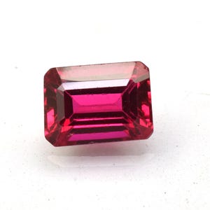 Könnte beinhalten: Ein leuchtender, tief pink-roter Edelstein im rechteckigen Smaragdschliff. Der Stein weist mehrere Facetten auf, die das Licht reflektieren und ein brillantes, durchscheinendes Aussehen mit scharfen, sauberen Kanten und einer glatten Oberfläche erzeugen.