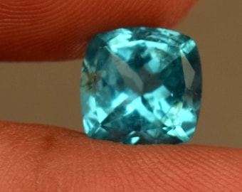 May include: A cushion-cut, aquamarine gemstone held between fingers. The gemstone is a vibrant, light blue color with multiple facets that reflect light. The gem is square with rounded corners.