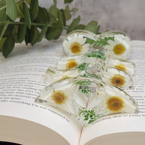 May include: A set of four clear resin hair clips with pressed white daisies and green leaves. The clips are stacked on top of each other.