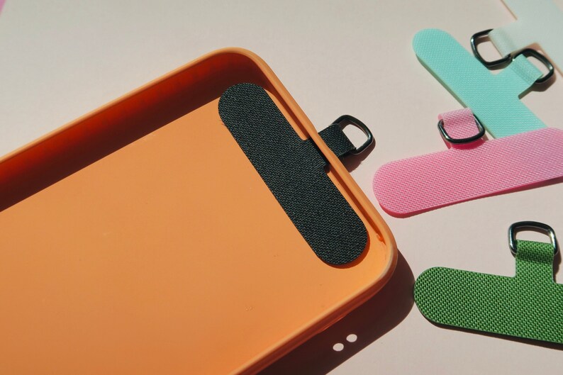 May include: An orange phone case with a black adhesive strap. The strap has a metal ring for attaching a lanyard or other accessories. The case is shown on a pink surface.