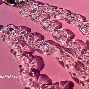 May include: A close-up of a clear plastic chain necklace with silver clasps. The chain is thick and chunky, and the links are large and oval-shaped. The necklace is lying on a pink surface.