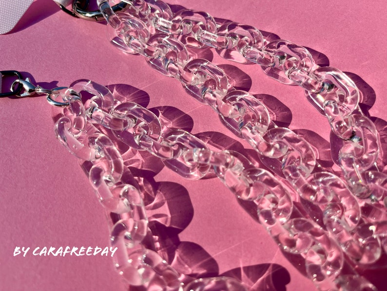 May include: A close-up of a clear plastic chain necklace with silver clasps. The chain is thick and chunky, and the links are large and oval-shaped. The necklace is lying on a pink surface.