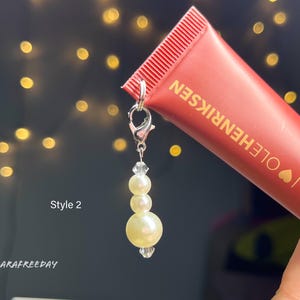 May include: A silver charm with a string of white pearls attached to a tube of lip treatment. The tube is red and has the text "I LOVE HENRIKSEN" on it. The text "Pout Peptide Lip Treatment" is also visible on the tube. The charm is labeled "Style 2".