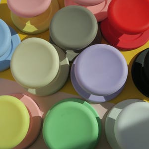 May include: A selection of pastel-coloured phone grips, including blue, pink, green, and yellow. The circular grips are displayed on a yellow and white surface, creating a visually appealing arrangement.