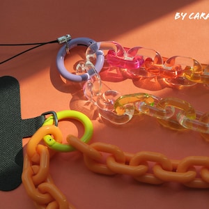 Puede incluir: Tres cadenas de plástico de colores con una correa negra y un anillo morado. Las cadenas están hechas de plástico transparente y son de los colores del arcoíris. Las cadenas están conectadas a una correa negra con un anillo morado. La correa tiene un pequeño lazo negro en la parte superior. BY CARAFREEDAY está escrito en blanco sobre un fondo negro.