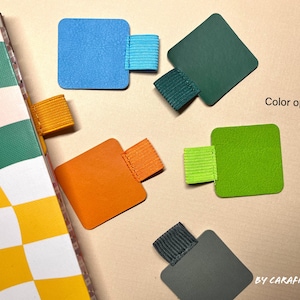 Può includere: Assortimento di segnalibri quadrati di diversi colori con bande elastiche. I colori includono blu, verde, arancione, giallo e grigio. Il testo "Color options" è visibile nell'immagine.