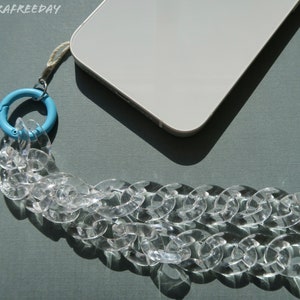 Op de afbeelding: Een transparante telefoonriem van kunststofketting met een lichtblauwe draaibare sluiting. De riem is bevestigd aan een witte smartphone.