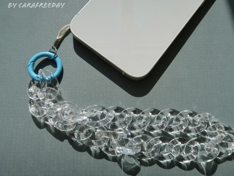 Op de afbeelding: Een transparante telefoonriem van kunststofketting met een lichtblauwe draaibare sluiting. De riem is bevestigd aan een witte smartphone.