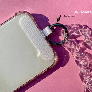 May include: A clear phone case with a white phone screen protector and a clear plastic chain with a metal ring attached. The chain is attached to the case with a white strap. The case and chain are on a pink surface.