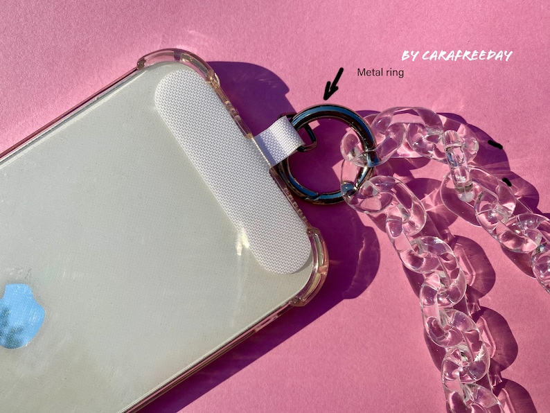 May include: A clear phone case with a white phone screen protector and a clear plastic chain with a metal ring attached. The chain is attached to the case with a white strap. The case and chain are on a pink surface.