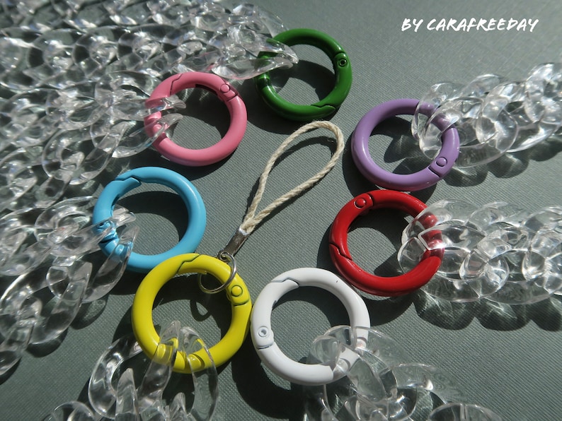 Op de afbeelding: Een transparante plastic ketting met zeven gekleurde metalen sluitingen. De sluitingen zijn in de kleuren roze, groen, paars, blauw, geel, rood en wit.