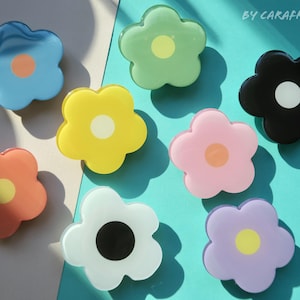 May include: A collection of eight flower-shaped accessories in various colors, including blue, green, yellow, black, pink, white, orange, and purple. Each flower has a contrasting center. The text "BY CARAFREEDAY" is visible in the upper right corner.