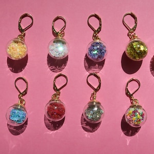 May include: A collection of gold-toned leverback earrings. Each earring features a clear glass orb filled with colorful glitter in various shapes and colors, including stars and confetti. The earrings are displayed on a pink background.