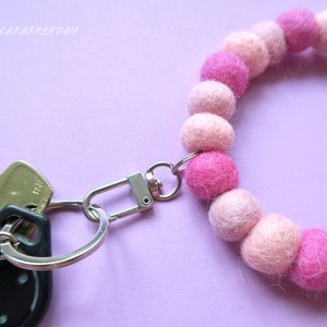 May include: A pink and white felt ball keychain with a silver key ring and a silver clasp. The keychain is attached to a set of keys.