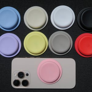 Peut inclure: Une collection de huit supports de téléphone circulaires au fini mat dans diverses couleurs unies, notamment bleu, beige, blanc, noir, violet, jaune, gris et rouge, avec un support rose fixé au dos d'un smartphone gris clair à trois caméras.