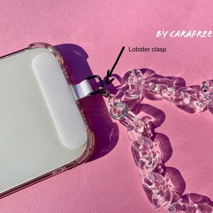 May include: A clear plastic phone chain with a lobster clasp attached to a white phone case. The chain is made of thick, chunky links. The phone case is clear and has a white border. The text "BY CARAFREEDAY" is visible in the top right corner of the image.