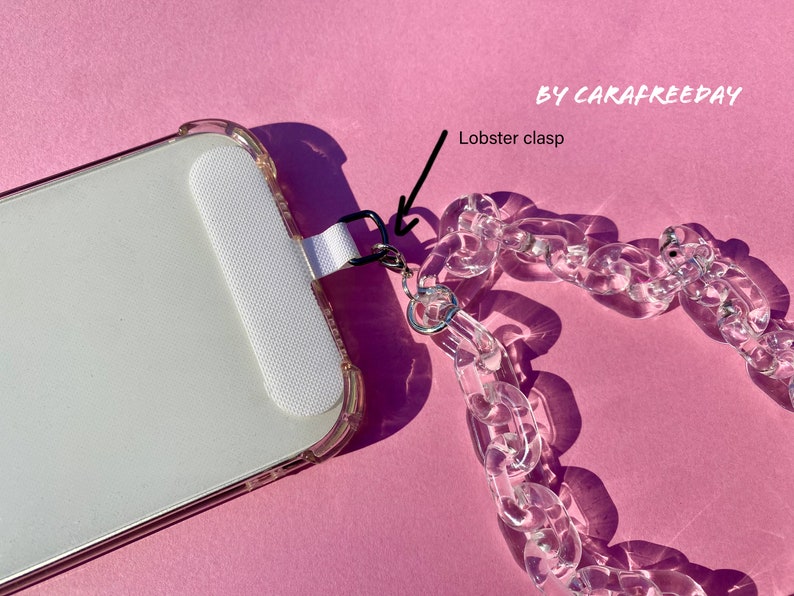 May include: A clear plastic phone chain with a lobster clasp attached to a white phone case. The chain is made of thick, chunky links. The phone case is clear and has a white border. The text "BY CARAFREEDAY" is visible in the top right corner of the image.