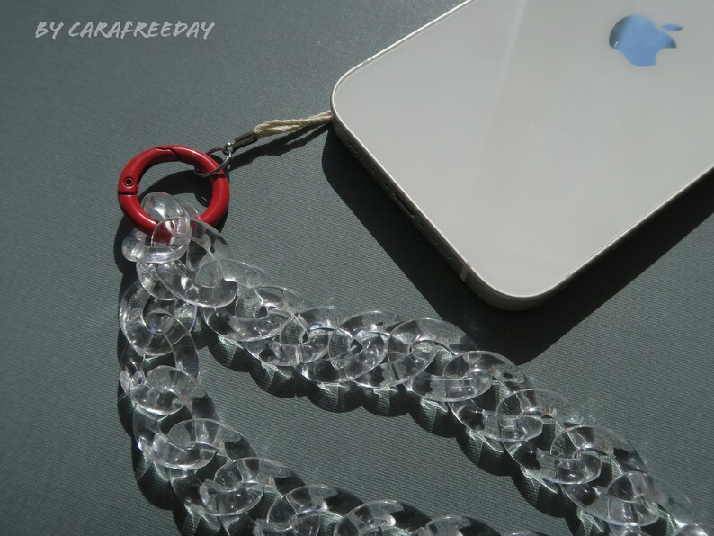 Op de afbeelding: Een transparante plastic kettingtelefoonband met een rode metalen sluiting. De band is bevestigd aan een witte smartphone met een zilveren Apple-logo.