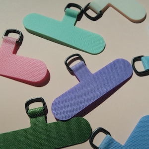 Op de afbeelding: Een verzameling kleurrijke telefoonbedels in roze, groen, blauw en paars. Elke bedel heeft een getextureerd oppervlak en een zilverkleurige metalen ring voor bevestiging. De bedels zijn gerangschikt op een lichtroze achtergrond.