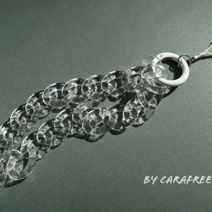Op de afbeelding: Een transparante plastic ketting telefoonbedel met een witte ring en een witte lus. De bedel is op een groene achtergrond.