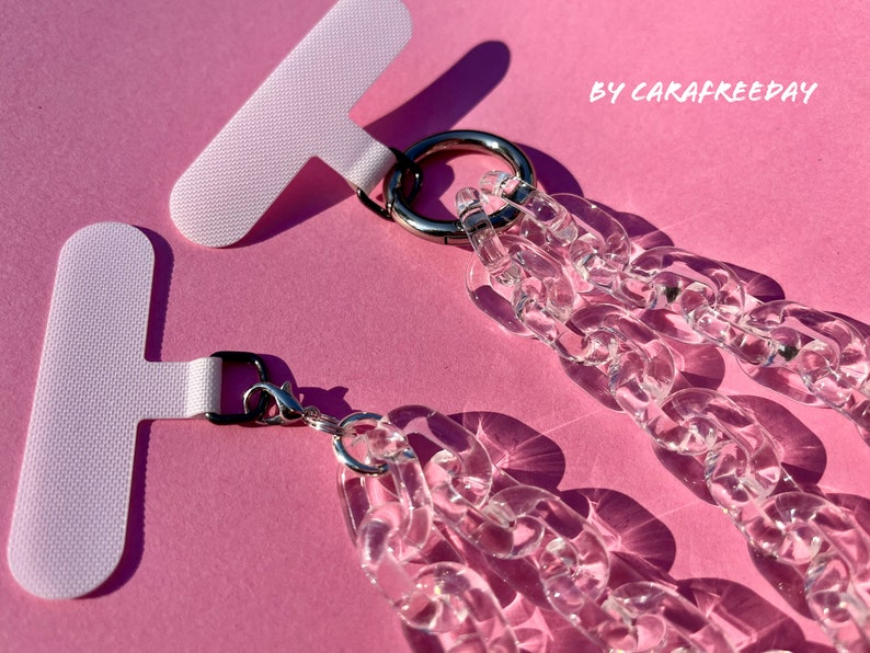 May include: Two clear acrylic chain phone straps with silver clasps and metal rings. The straps are attached to white T-shaped adhesive strips. The text "BY CARAFREEDAY" is visible in the upper right corner of the image.