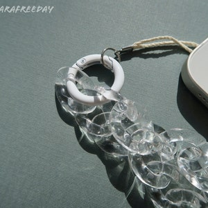 Op de afbeelding: Een transparante telefoonriem van kunststofketting met een witte metalen sluiting en een wit touwtje.