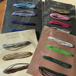 May include: Four sets of journal ribbons on rectangular cards, each featuring four distinct colored elastic cords. The cards display the text Journal Ribbons and 4 colors 3x45cm each. The cords come in blue, purple, pink, red, grey, tan, brown, green, and black.