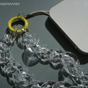 Op de afbeelding: Een transparante plastic ketting met een gele ring bevestigd aan een witte telefoonhoes. De ketting is gemaakt van grote, dikke schakels.