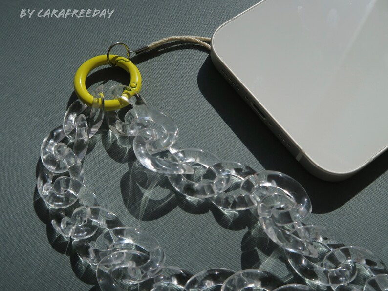 Op de afbeelding: Een transparante plastic ketting met een gele ring bevestigd aan een witte telefoonhoes. De ketting is gemaakt van grote, dikke schakels.